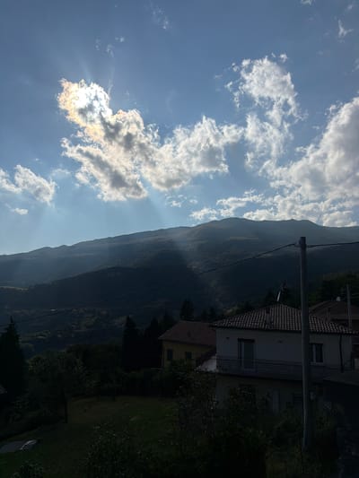 The image captures a landscape scene with a prominent mountain range silhouetted against a bright, partly cloudy sky. Sunlight filters through a cluster of clouds, creating visible sunbeams that descend towards the mountains. In the foreground, there are houses with terracotta roofs and a visible utility pole with wires. The lower part of the image is dominated by dark vegetation, suggesting a natural, possibly rural, setting. While the specific location is stated as Ferrara di Monte Baldo, Italy, the image itself provides no explicit visual cues that confirm this city. There are no people or animals visible in the frame. The scene appears to be an outdoor view, likely taken during daylight hours, with the sun positioned towards the left of the frame.