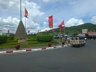 In Liên Nghĩa, Vietnam, a conical monument, adorned with a lattice of what appears to be recycled materials, stands prominently in a roundabout under a partly cloudy sky. The monument is surrounded by meticulously manicured green hedges and gardens, with large planters filled with pink flowers at the base of the roundabout's curb. A row of red flags, some featuring a yellow star, flutter in the breeze from tall poles surrounding the monument and throughout the park area.

To the right of the monument, a white utility truck with a covered bed, typical for transporting goods or passengers, drives away from the camera on the asphalt road. The truck has a license plate that reads "49H-016.74". Beyond the roundabout, buildings with commercial signage are visible, suggesting a developed urban area. In the background, a lush green, tree-covered mountain range forms the backdrop to the scene. The road is clearly marked with a red and white striped curb.