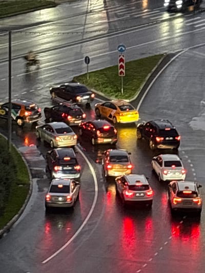 This aerial-view, low-light photograph captures a traffic jam on a wet, paved road, likely in Лесные Поляны, Russia. The scene is dominated by numerous cars, some with their brake lights illuminated, suggesting the vehicles are stationary or moving slowly. The road appears to be a multi-lane intersection or a stretch of highway with a curve.
The time of day is dusk or night, as indicated by the low illumination and the glowing lights of the vehicles. The wet surface of the road reflects the lights, enhancing the moody atmosphere. A patch of green grass is visible to the left of the road, with a traffic sign featuring a blue circle with a white arrow and a red and white directional arrow sign to its right, which suggests a turn or lane guidance.
The cars are a mix of colors and models, but their details are somewhat obscured by the lighting conditions and the perspective. There are no discernible people visible in the cars or on the road. The overall impression is one of stalled progress and a somber, perhaps frustrating, moment for the drivers caught in the congestion. The weather appears to be damp, contributing to the reflections on the road surface.