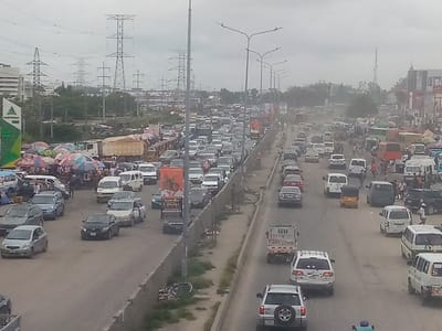 The image displays a heavily congested urban street scene in Ajah, Nigeria, under an overcast sky. A multi-lane dual carriageway is visible, with dense vehicular traffic in both directions appearing stationary or moving slowly. Vehicles include numerous sedans, SUVs, white vans, a large beige dump truck, and a yellow tricycle ("keke Napep").
On the left side of the road, a vibrant street market is set up, featuring multiple vendors operating from stalls shaded by colorful, striped umbrellas (red, blue, green, yellow). Numerous pedestrians are visible congregating around these market stalls and moving alongside the road. A prominent green sign with "NORTHWEST" is partially visible on the far left, indicating a commercial establishment.
Along the central divider of the road, tall poles support multi-armed streetlights. Further into the background, a series of large electricity transmission towers (pylons) extends across the landscape. Commercial buildings, including multi-story structures, are present on both sides of the thoroughfare, along with large advertising billboards. A discernible haze or dust is present in the distant air.