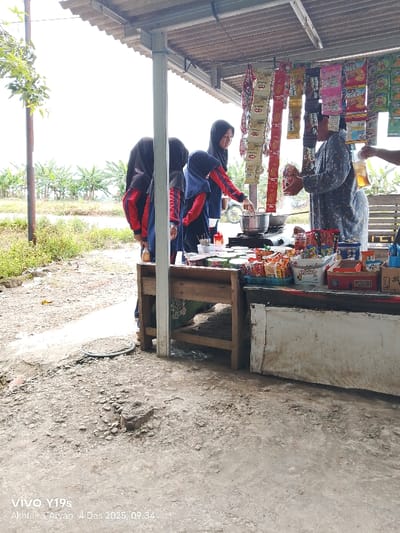 Four young women wearing hijabs and matching red and blue jackets stand behind a wooden counter. A person in a grey patterned shirt is pouring liquid into a clear cup. Snacks and various items hang above the counter. The items are arranged neatly. The ground is rough and uneven. Banana trees line the horizon. A date stamp at the bottom left of the image reads "VIVO Y19s Akhtar & Aryan 4 Des 2025, 09:34".