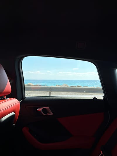 The image is a view from inside a car, looking out the rear passenger window. The car's interior features red leather seats and black door panels with a silver door handle. Outside the window, a bright blue ocean is visible under a clear sky with scattered white clouds. In the middle ground, there is a rocky shoreline and a wooden bench. A faint line of what appears to be a railing runs along the shoreline. The sun is shining brightly, casting strong shadows within the car. The scene depicts a scenic coastal drive in Kissonerga Community, Cyprus.