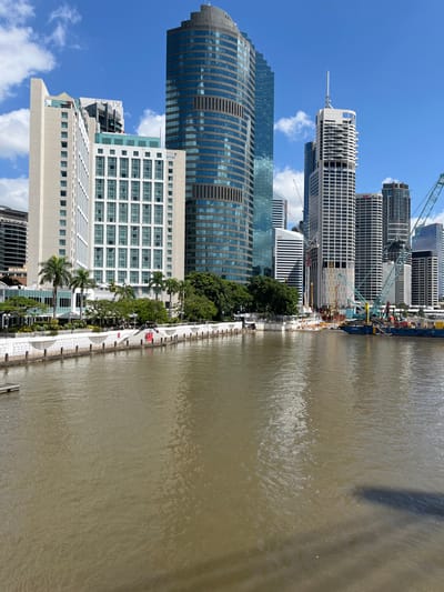 This image presents a vibrant urban landscape along the Brisbane River in Brisbane City, Australia, under a clear blue sky dotted with fluffy white clouds. The scene is captured from an elevated perspective, looking down on the river and its adjacent city skyline.

On the left, a dense collection of modern high-rise buildings dominates the foreground and midground. A distinctive, tall building with a curved façade and blue reflective glass stands prominently. Alongside it, numerous other skyscrapers, some exhibiting intricate window patterns and lighter exteriors, rise towards the sky. Towards the middle and right of the building cluster, several structures are clearly under construction, indicated by the presence of multiple large cranes that extend above the developing edifices.

The center of the frame features a well-designed riverside promenade. This wide, paved walkway displays a decorative pattern and is bordered by a continuous line of palm trees and other green foliage, providing a lush urban green space. A few small, indistinct figures are visible on the promenade, including a notable red element, possibly a person or a small recreational vehicle, adding a sense of activity. Bollards line the edge of the path closest to the water.

The right side of the image is filled by the broad, brownish-green expanse of the Brisbane River. Its surface shows gentle ripples, reflecting the bright daylight. Long shadows from tall structures fall across the water, suggesting the time is likely late morning or early afternoon. A work vessel or barge is docked near the construction area on the river, indicating ongoing urban development or maintenance activities. The overall impression is one of a bustling, modern city enjoying sunny weather. No discernible text is visible in the image.