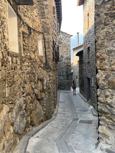The image depicts a narrow, cobblestone alleyway in the historic district of Benasque, Spain, with stone buildings lining both sides. A young woman with a red backpack is walking away from the camera down the alley. The buildings are constructed from rough-hewn stone, with some featuring wooden shutters and windows. Electrical wires are visible running along the facade of one building. The lighting suggests it is late afternoon, casting shadows down the alley. The overall scene conveys a sense of rustic charm and historical preservation.