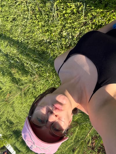 The image shows a person lying on the grass, possibly in an outdoor setting like a park or backyard. The person is wearing a pink baseball cap and glasses. They appear to be looking up at the camera. The background consists of lush green grass and some foliage. The lighting suggests it's a sunny day. There is no specific information visible in the image to determine the location within the city of Докша, Russia.