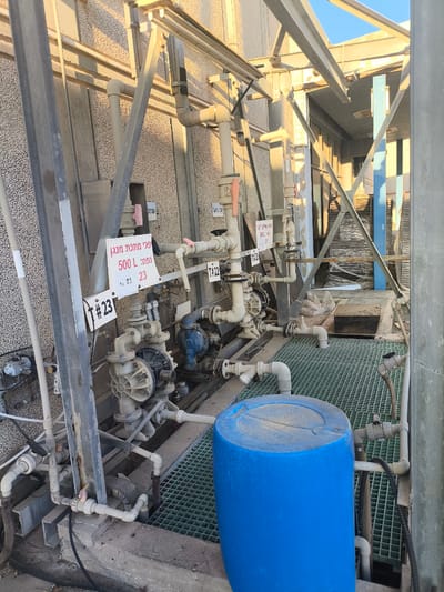 The image shows an industrial setting, likely a chemical processing plant, located in Dimona, Israel. The scene is dominated by a complex network of pipes, pumps, and valves, suggesting an active or recently active industrial process. A large blue plastic drum sits on a green metal grating, which appears to be part of a secondary containment system. The lighting suggests it is daytime, with sunlight casting distinct shadows, indicating a sunny weather condition. The overall impression is one of a functional, albeit somewhat aged, industrial environment.
Several signs are visible, providing text in Hebrew. One sign reads "שפכים אתילן גליקול" (Ethylene Glycol Wastewater), and another states "שפכי מתכת מנגן" (Manganese Metal Wastewater), identifying the types of materials handled or processed. There are also labels with numbers, such as "T#22" and "#23", likely for identification or asset management. The presence of these labels and the industrial infrastructure points to a specific operational area within the facility. No people are visible in the frame, but the equipment suggests ongoing or potential activity. The setting appears to be outdoors or in a covered open-air structure, given the sunlight and the type of construction materials visible, such as corrugated metal siding and steel beams.