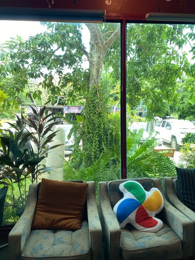 The image shows an indoor seating area looking out through a large window. In the foreground, two armchairs are visible, each with a cushion. The left armchair has a brown rectangular cushion. The right armchair has a large, white, multi-colored pillow shaped like a pinwheel. The armchairs have a light-colored fabric with a blue floral pattern. Outside the window, lush green foliage and trees dominate the scene. A large tree trunk is prominent, covered in hanging vines. In the background, a white pickup truck is parked, and a sign that reads "FOOD" in Thai script is visible. The overall environment suggests a relaxed outdoor setting, possibly a cafe or restaurant in Bang Sare, Thailand.