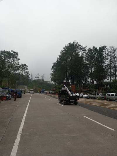 The image shows a road scene in Mae Chan District, Thailand, characterized by an overcast sky. The road, marked with white lines, has various vehicles present including a black pickup truck carrying metal poles, a white van, and several smaller cars. On the side of the road, there are shops, trees, and some parked vehicles, including a motorcycle. People are visible near the shops. Further up the road there is a bridge. The environment includes trees and hills.