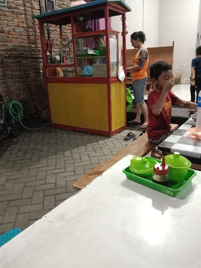 A young boy is sitting at a table in front of a food stall in Kecamatan Brangsong, Indonesia. He is wearing a red shirt and blue shorts, and is looking towards the camera with his mouth slightly open. There are two green condiment containers on the table in front of him, along with a brown sauce dispenser. The table is made of white laminate and has a wooden edge. Behind the boy, an adult female is standing in front of the food stall, wearing a yellow shirt and blue denim shorts. The stall is a yellow and red structure with shelves and a glass front. The stall is set up on a brick patio. The patio is made of gray bricks arranged in a herringbone pattern. The background of the image is a brick wall. There is a black motorcycle parked in the background. The image is taken from an angle looking down at the table.