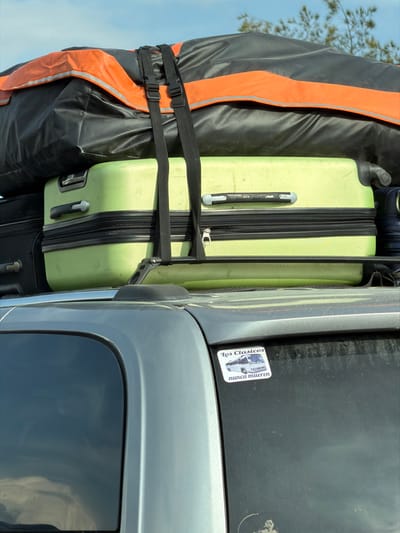 Luggage secured on top of a vehicle includes a black duffel with orange accents and two suitcases, one light green and another dark. Black straps fasten the luggage. A sticker on the vehicle's roof reads "Los Clasicos nunca mueren" with a vintage car graphic. The car is silver, with a dark window reflecting the sky.
