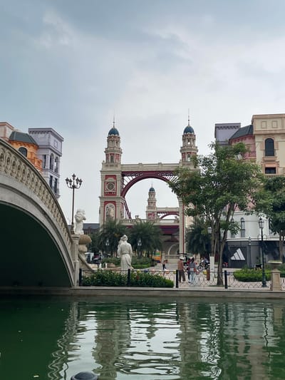 The image depicts a canal in front of a European-inspired architectural complex in the city of Nghĩa Trụ Commune, Vietnam. The canal is filled with murky green water that reflects the surrounding structures. On the left, a large, ornate arched bridge made of light-colored stone dominates the foreground. The bridge's railing is decorated with a continuous pattern of circles.
Beyond the bridge, on the right side of the canal, a pedestrian walkway is visible. Several people are present on the walkway. Two women and a man are walking together, dressed in casual attire. Another person is seen cycling. In the background, multiple buildings with distinct architectural styles are visible. These buildings feature colorful facades in shades of purple, pink, and beige, with elements like towers, domes, and balconies. One building has an Adidas logo displayed.
The central focus of the background is a large, red arch bridge with two prominent towers on either side. These towers have ornate spires and clock faces. Further in the distance, another similar tower can be seen through the arch of the red bridge. Palm trees and other greenery are interspersed among the buildings. The sky is overcast, with a muted, grayish-blue hue. Two white statues of human figures are positioned near the bridge and walkway. The overall scene suggests a theme park or a meticulously designed public space.
