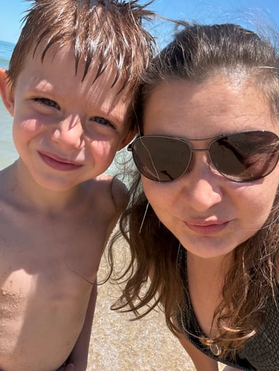 This media file is a close-up, selfie-style photograph featuring two individuals at a beach, likely Shkorpilovtsi, Bulgaria. The subjects are a woman and a young boy, positioned in a way that suggests they are taking the photo together. The woman, wearing sunglasses, is in the upper portion of the frame, her face partially obscured by the lenses and the bright sunlight. Her long, brown hair is visible, some strands falling across her face and shoulders. The boy is in the foreground, his head tilted upwards towards the camera. He has wet, brown hair and is smiling, with his eyes looking directly at the viewer.
The setting is a beach, indicated by the presence of sand, water, and the bright, sunny atmosphere. The water appears to be shallow, with gentle ripples visible in the background. The lighting suggests it is daytime, likely late morning or early afternoon, given the strong shadows and highlights on the subjects' skin. The overall emotion conveyed is one of happiness and enjoyment, typical of a day at the beach. The woman's expression is partially hidden, but the boy's smile is clear and joyful. The image captures a moment of connection and fun between the two individuals in a natural, outdoor environment. There is no visible text in the media file.