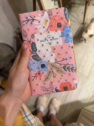 A person's hand holds a pink notebook adorned with a floral pattern and glitter accents. The notebook has a white label with the text "NOTE BOOK" and "FLOWER". In the background, a blurred white and tan cat is visible, along with feet wearing fluffy slippers and white socks. The scene appears to be indoors, with a wooden floor and a glimpse of furniture. There is no discernible connection to the city of Semiluki, Russia.