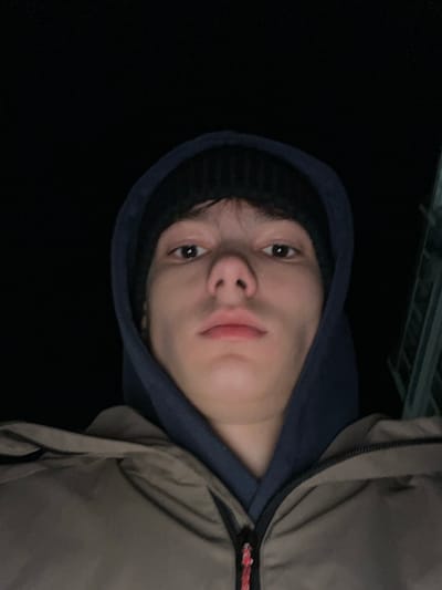 An upward-angle selfie depicts a single young male against a predominantly dark background, suggesting nighttime or low light conditions in Zainsk, Russia. The male has fair skin, dark brown eyes, and a neutral expression. He is wearing a black knitted beanie, a dark blue hooded sweatshirt with the hood pulled up, and a khaki or olive-green puffer jacket. The jacket features a zipper with a red, white, and black cord pull. A faint, light-colored structure with vertical and horizontal elements is partially visible in the upper right background. The subject's face is illuminated from the front, likely by a flash or screen light.