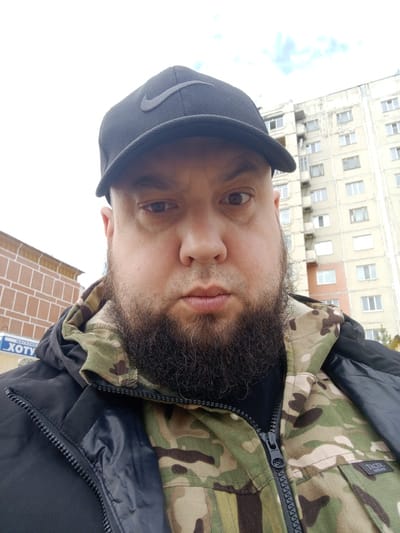 A man with a dark beard and a black Nike cap is the central subject of the image. He is wearing a green camouflage jacket over a black jacket. The zip of the camo jacket is partially visible. Behind the man, on the left side of the frame, there is a building constructed from red bricks with what appears to be Cyrillic writing. To the right of the man, a multistory apartment building is visible. The building has multiple windows. The sky is overcast. The location is Chulman, Russia.
