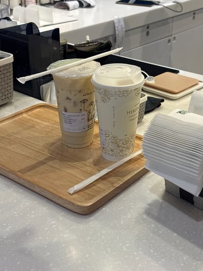 The image captures a close-up view of a counter in a beverage establishment, likely in Nong Pa Khrang, Thailand. Two takeaway cups are prominently displayed on a wooden tray. The cup on the left, a clear plastic one, contains a light-colored milky beverage with ice, and its label indicates "Camellia Milk Tea L, Sugar 25% (1 P), Original Milk." A white paper straw is resting on its lid. The cup on the right is made of cream-colored paper, adorned with floral patterns and text that appears to be in Japanese and English, reading "KAHO HERITAGE TEAFLOWER." Another white paper straw is positioned on its lid. To the right of the cups, a stack of folded white paper napkins is visible in a grey woven basket. The counter surface is light grey with a speckled texture. In the background, a black dispenser unit and other indistinct objects are partially visible. The overall lighting suggests an indoor environment.