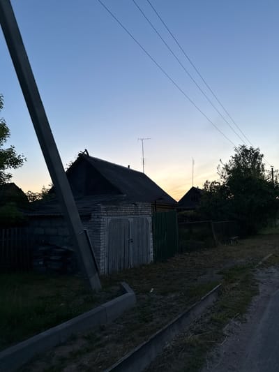 The image captures a tranquil, rural or semi-rural scene, likely in Лабжанскі сельскі Савет, Belarus, during either sunrise or sunset. The sky transitions from a warm, golden glow near the horizon to a pale blue overhead, indicative of twilight hours. There are no clouds, suggesting clear weather.
In the mid-ground, a prominent, rustic building stands with a dark, sloped roof, brick walls, and a visible wooden door, suggesting it might be a barn, garage, or an older dwelling. Behind this structure, another building is partially visible. Two antennas are mounted on poles, one on the main building's roof and another further back, implying communication or broadcast reception.
Power lines stretch across the upper portion of the sky, adding an element of infrastructure to the natural setting. Dark green trees and dense foliage frame the buildings and parts of the sky, creating a contrast with the glowing horizon.
In the foreground, a dark, possibly metal or concrete, barrier or structure runs diagonally across the lower part of the frame. To the right, a dirt or unpaved path is visible, bordered by grass and weeds, alongside a concrete or stone-lined drainage channel.
The scene is static, with no discernible human activity. The overall impression is one of quietude and the simple beauty of a countryside evening or morning. No visible text is present in the image.
