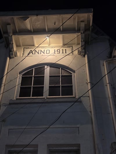 The image shows the facade of an old building at night, illuminated by a spotlight. The building has a sign that reads "ANNO 1911" above a large arched window with multiple panes. Several dark power lines are strung across the facade. The building appears to be made of light-colored brick or stucco, with some decorative architectural elements. The overall impression is one of age and historical significance.