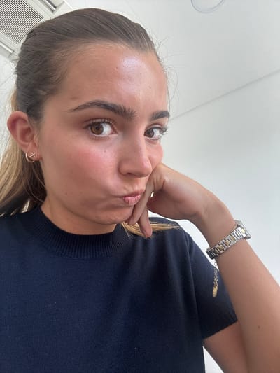 A light-skinned woman with brown hair and eyes is visible in a close-up, slightly distorted photograph, taken from a low angle. She is wearing a dark blue sweater and a silver and gold wristwatch on her left wrist. She is holding her hand to her face, resting her fingers against her cheek and lips. The background is a plain white wall. The woman has a small gold cross earring in her left ear. The photograph does not contain any location information about Torres Vedras, Portugal.