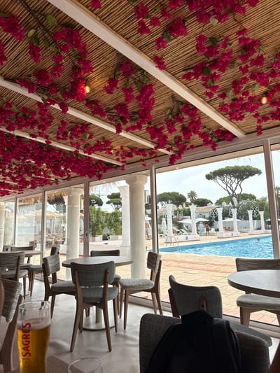 This image presents a view from an indoor dining or lounge area looking out onto a swimming pool and resort grounds in Almancil, Portugal, identified as The Magnolia Hotel in Quinta do Lago. The indoor ceiling is covered with a bamboo-slat canopy from which numerous strands of artificial fuchsia bougainvillea flowers descend, illuminated by small, exposed light bulbs. White structural beams are visible. The foreground features multiple round tables with grey tops and chairs with grey fabric upholstery and light wooden frames. On a table in the lower left, a glass of light-colored liquid, displaying "SAGRES" branding, is present alongside an empty glass. A dark garment is draped over a chair in the lower right.

Large glass panels, supported by white classical columns, separate the indoor space from the outdoor area. Outside, a blue swimming pool is visible, bordered by a light-colored paved deck with reddish-brown stripes. Several individuals are present in the outdoor scene: one person is in the pool, others are reclining on white sunbeds under white umbrellas, and some are walking. The poolside area is adorned with additional white classical columns and distinctive white pillars featuring stylized wolf heads. The background includes extensive green vegetation, primarily large umbrella pines, and partially visible white buildings with terracotta roofs under a partly cloudy sky.
