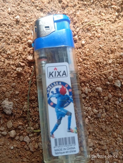 A close-up shot displays a blue and silver cigarette lighter resting on sandy ground. The lighter features a clear plastic body with a blue top and a silver metal wheel. A label on the lighter shows the brand name "KIXA" and an image of a soccer player in a blue uniform with a red flame on his head, along with the words "CHELSEA" and "BALL". Below the image, a barcode is visible, followed by the text "MADE IN CHINA" and "fabrique en chine". In the lower right corner, a timestamp reads "11/06/2026 09:05". The ground beneath the lighter is composed of loose sand and small pebbles, with a few dry blades of grass scattered around. The lighting suggests it is daytime, with the sun casting a slight glare on the lighter. The specific location in DU, Nigeria, is not identifiable from the image.