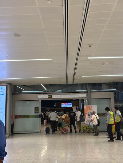 In Gollapalle Khurd, India, an indoor airport scene is captured, focusing on a group of people near an exit. The ceiling is a modern, white, tiled structure with integrated lighting and circular fixtures. In the foreground, the polished floor reflects the ambient light.

A sign with the word "Exit" is prominently displayed above an entryway. To the left, a large digital advertisement screen is partially visible. Several individuals are walking, some with luggage. One person is pulling a dark suitcase on wheels, while another walks with a backpack. Two men in high-visibility vests, possibly airport staff, are walking together on the right side of the frame. In the background, a banner for the "11th All India Police Judo Cluster 2026" is visible. The overall environment suggests a busy transportation hub.