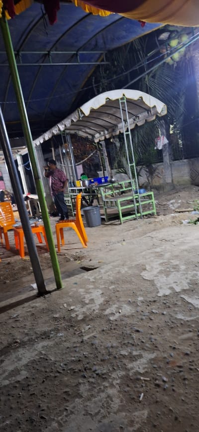This is a full-frame, eye-level, vertical shot of an outdoor area in Payaterbang, Indonesia. The scene appears to be a temporary seating area, possibly for an event or a food stall, under a large, blue tarpaulin roof. In the foreground, an orange plastic chair is visible on the left, and further back, another orange plastic chair is positioned. A man in a patterned shirt and dark pants stands on an orange plastic chair, holding a cell phone to his ear. He is the primary human subject.

Behind the man, there is a table with some items on it, including blue containers and what looks like a green container. To the right of the table, there is a green metal frame structure with shelves, possibly for seating or display. A grey plastic trash can is located near the base of this structure. The ground is rough, uneven, and appears to be dirt or gravel with some debris. In the background, there are trees and some structures, suggesting a built-up area. The lighting is dim, indicating it might be evening or nighttime. The overall environment suggests a casual, possibly temporary, gathering space.