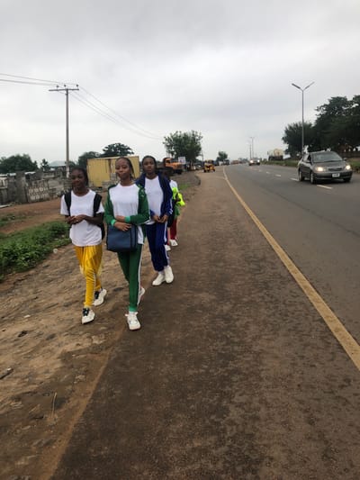 The image shows a group of four young women in tracksuits lying in a line on the edge of a paved road. They are wearing white shirts and different colored track pants: yellow, green, and blue. The road has a yellow dividing line. Cars are visible in the distance, moving along the road. There is a building under construction nearby. Trees and electrical poles are visible in the background. The sky is overcast. The image was captured in the city of Karu, Nigeria.
