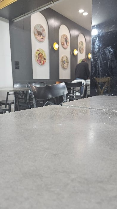 A sideways shot reveals a restaurant interior. Dark grey walls are adorned with decorative plates and illuminated by recessed lighting. Seating includes black chairs and tables, partially occupied by a person wearing dark clothing. The foreground features a grey textured surface, possibly a floor or wall, creating a contrast with the darker interior. The image is slightly out of focus, giving it a soft, almost blurry appearance.