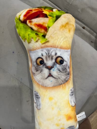 The image shows a stuffed toy shaped like a burrito with a cat's face printed on it. The toy is light-colored, resembling a tortilla, with colorful filling visible at the top, including green lettuce, red sauce, and white meat. The cat's face is centrally located on the toy, featuring large, surprised eyes, gray and white fur markings, and a small pink mouth. Two gray cat paws are depicted on either side of the cat's face. The toy is lying on a textured gray surface, likely upholstery, possibly a couch or chair. A small white tag with laundry symbols and Russian text is attached to the lower right side of the toy. The background is blurred and out of focus, showing a continuation of the gray surface and hints of wood grain on the floor. There is no visible indication of the city of Gorodets, Russia, within the image.