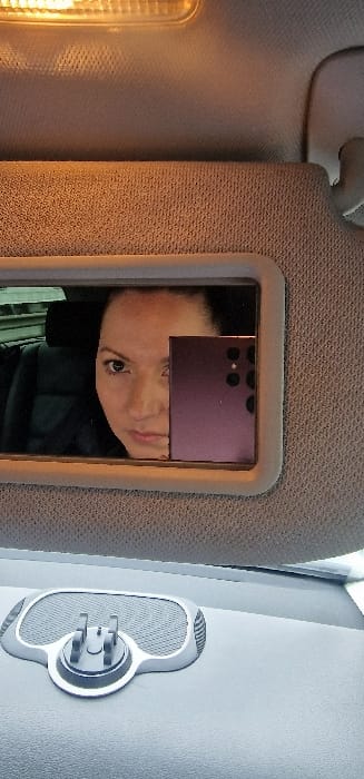 The scene depicts the interior of a vehicle, located in Cenon, France. A beige fabric sun visor, partially lowered, is positioned in the upper half of the frame, containing a rectangular vanity mirror. Reflected within this mirror is a female individual with dark hair, looking directly forward. Her face is partially obscured on the right side by a purple smartphone she is holding, which features multiple rear camera lenses and is being used to capture the image. Dark car interior elements, including seating, are visible behind the reflected individual. An illuminated yellow-orange overhead light fixture is present at the top of the frame. The lower portion of the image reveals a dark gray dashboard, with an attached, empty gray and black smartphone mount situated on the left side.
