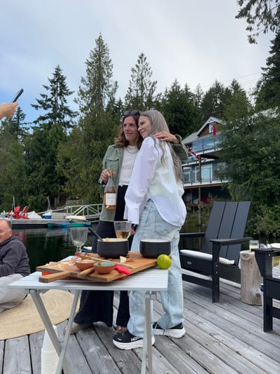 The image depicts an outdoor social gathering on a wooden dock at Area B (Shawnigan Lake), Canada, under an overcast sky. Two adult females are positioned centrally, one with brown hair wearing a green jacket over a white sweater and black pants, holding a bottle of rosé wine. The other, with long grey hair, wears a white collared shirt over light blue jeans and black and white Hoka shoes, with her left arm around the first woman. Both are smiling and looking towards the left. A partially visible hand holding a black smartphone is visible in the upper left corner, aimed towards the two women.

In the foreground, a silver collapsible table holds a light brown wooden serving board. The board features a variety of small bowls containing food items, a whole lime, and a red-handled knife. Two clear wine glasses are also present on the table, one containing a light-colored liquid. To the left of the table, a partially visible adult male with short dark hair is seated. A circular, light-brown woven mat is visible beneath the table, and a stack of white disposable cups rests on the dock surface.

In the background, calm, dark lake water extends behind the dock. Several small boats, including kayaks and paddleboards, are moored near the dock. Tall evergreen trees line the distant shore. A multi-level house with grey siding, brown accents, glass railings, and a prominent red Canadian flag hanging from a balcony is visible amongst the trees on the opposite shore or a nearby property. Two black Adirondack chairs and a wooden stump table are visible to the right of the central figures on the dock.