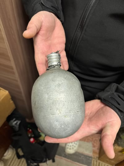 A person wearing a black jacket is holding a metal canteen. The canteen is cylindrical with a rounded body and a threaded neck with a metal cap attached by a chain. The hands holding the canteen are pale. The background is slightly blurred but shows a wooden surface and indistinct objects. The location is not discernible from the image and the query about Маза, Russia is irrelevant.