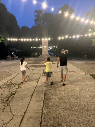 In Parvomay, Bulgaria, at dusk, a man, a young boy, and a young girl walk away from the camera on a concrete path in a park. The man is wearing a black t-shirt and light-colored shorts. The boy is wearing a yellow t-shirt and striped shorts. The girl is wearing a white t-shirt and grey shorts. A small brown dog is walking in front of the boy. In the background, there is a monument with a star on top, and rows of string lights are hung overhead. Trees line the sides of the park, and there are benches and a small building visible in the distance. The sky is dark blue, indicating it is twilight. The ground has cracked concrete sections.