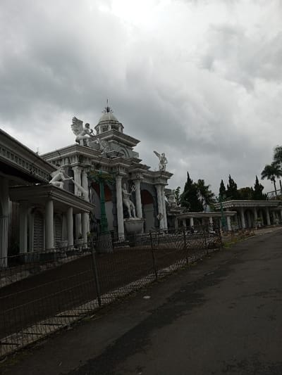 An exterior view depicts a large, ornate neoclassical-style building facade or gate located in Melakudikadu, India, under a heavily overcast, light grey sky. The structure, predominantly light grey in color, features multiple classical columns, arched recesses, intricate cornices, and a central dome surmounted by a finial. Numerous white-colored statues are integrated into the architecture: a winged figure stands centrally on the uppermost roof, another figure with a raised arm is positioned on the right roofline, a crouching figure occupies a lower left roofline, and a male figure holding a trident stands at ground level on the right side of the main entryway. Dark green, ornate lampposts are visible within the facade. A dark asphalt road, appearing somewhat textured, runs across the foreground and curves along the right, separated from the building's base by a low, dark chain-link fence. Behind the edifice, a line of slender coniferous trees and several prominent palm trees are visible. No human subjects or discernible actions are present in the static scene.