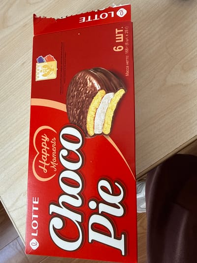 The scene is set indoors, depicted as being in the city of Aragats, Armenia. A red Lotte Choco Pie box lies on a light-colored wooden surface, possibly a table or desk, oriented vertically with its top facing the left side of the frame. The box features an image of a cut Choco Pie revealing its chocolate coating, two layers of yellow cake, and a white cream filling. Prominently displayed text includes "LOTTE," "Happy Moments," and "Choco Pie" in large white letters. Other text visible in Russian states "6 ШТ." (6 pcs) and "Масса нетто: 168 г (6 шт. х 28 г)" (Net weight: 168g (6 pcs x 28g)), along with "ОФИЦИАЛЬНЫЙ ПОСТАВЩИК МОСКОВСКОГО КРЕМЛЯ" (Official supplier to the Moscow Kremlin) accompanied by a crest. The top edge of the box shows minor tearing and creasing, consistent with recent opening or handling. In the lower right corner, a portion of a person's leg, clad in dark reddish-brown trousers, and the front part of a light-colored athletic shoe are visible, positioned below the wooden surface. No other subjects or actions are discernible.