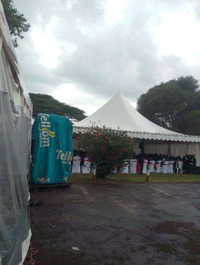 This image depicts an outdoor event venue in Gitaru, Kenya, set up under an overcast sky. The ground, a dark asphalt, appears wet, suggesting recent rainfall.
The scene is dominated by two white event tents. In the mid-ground, a large, rectangular tent features scalloped edges and provides shelter for numerous white chairs arranged in rows. Many of these chairs are decorated with red and blue sashes, hinting at a formal gathering or celebration. Behind this main structure, a smaller, conical white tent stands.
To the left, a tall, green object covered in a branded banner prominently displays the "Telkom" logo and the text "MOVING WITH YOU," indicating corporate involvement, likely a sponsorship or a company event. A vibrant bush with bright red flowers is situated in front of the main tent, flanked by two short, striped poles (red, white, and green).
In the background, lush green trees contribute to the outdoor setting. The visible seating arrangement suggests an organized gathering or ceremony, possibly a community or corporate event given the Telkom branding. The overall atmosphere is one of anticipation for or during an event, under grey, cloudy conditions.