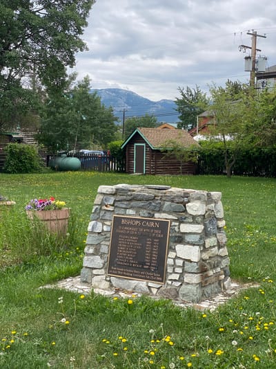 The image depicts an outdoor scene in Whitehorse, Canada, focused on a stone monument in a grassy area dotted with dandelions. The monument, constructed from various stones, features a bronze plaque inscribed with text. The plaque's title reads "BISHOPS CAIRN" and lists several names with corresponding years, seemingly indicating a historical or commemorative purpose. Below this list, it states, "From friends to honour Betty and Charles Taylor on their 50th Wedding Anniversary, Feb 23rd 1956."
In the background, a rustic wooden shed with a brown roof is visible, set against a backdrop of lush green trees and a chain-link fence. Beyond the fence, two large green tanks are situated, and a blue vehicle is parked behind them. A distant mountain range, partially obscured by clouds, forms the horizon. The sky is overcast, contributing to the soft, diffused lighting of the scene. The overall impression is one of a peaceful, possibly historic, location within the city, marked by the monument and its surrounding natural elements.