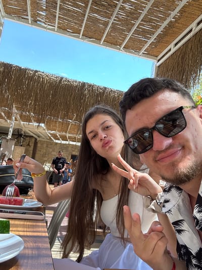 A selfie captures a man and a woman enjoying an outdoor meal on a bright, sunny day in Selçuk, Turkey. The image is oriented with the man and woman angled diagonally, suggesting a portrait-style selfie taken from above.
The man, positioned closer to the camera, has dark hair, a light beard, and is wearing black sunglasses that reflect the surroundings. He wears a white shirt with a black palm tree pattern. He is smiling slightly, looking towards the camera. The woman is positioned below and slightly to the side of the man, with long dark hair, wearing a white top. She is playfully pouting her lips and making a peace sign with her left hand. A yellow wristband with illegible text is visible on her right wrist.
They appear to be seated at a table, with parts of a meal visible, including slices of watermelon and cucumber on plates. A rose gold smartphone and a black handbag are also on the wooden table. The woman holds a fork with a piece of watermelon, suggesting she was eating or about to eat.
The background shows a clear blue sky and a distinctive thatched or bamboo roof structure providing shade, indicative of an outdoor restaurant or cafe setting. In the mid-ground, other patrons are visible seated at tables, and a stone wall structure is present. The overall atmosphere is relaxed and casual, characteristic of a holiday or leisure setting.