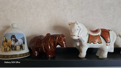 The image displays a shelf containing three decorative horse figurines. On the left is a bell-shaped figurine with a painting of two horses. To its right is a brown, glossy ceramic horse with a saddle. The furthest figurine on the right is a white, glossy ceramic horse adorned with a red and gold saddle blanket. The background is a textured, off-white wall. The shelf itself is black. The lower portion of the image shows a portion of the text "Galaxy S24 Ultra." The scene appears to be indoors. There is no information to suggest this is located in Khatassy, Russia.