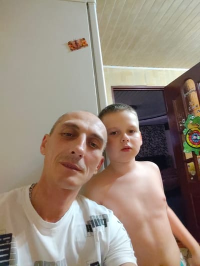 The image is a selfie taken indoors. Two subjects are visible: a man in the foreground and a young boy behind him. The man is bald and wearing a white t-shirt with a silver chain. He is looking at the camera with a neutral expression. The boy is shirtless and has short, light brown hair. He is also looking towards the camera with a slightly curious expression.
In the background, a refrigerator is partially visible on the left. A decorative magnet with an image of a butterfly is attached to the refrigerator door. To the right, a wooden door is visible, adorned with several colorful decorations, including a frog-shaped target. The room appears to be a home, possibly a kitchen or hallway, with a wood-paneled ceiling. The lighting suggests it is daytime. The overall mood is casual and familial. No visible text is discernible in the image beyond what might be on the t-shirt pattern.