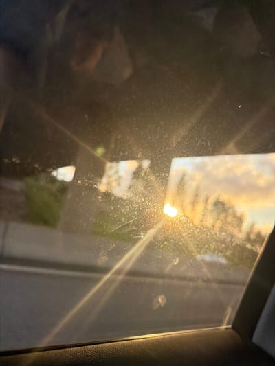 The image is a photograph taken from inside a vehicle, looking out through a dirty window. The sun is low in the sky, casting a warm, golden light that creates a lens flare across the image. In the distance, through the window, the sky is filled with soft, wispy clouds illuminated by the setting sun. Silhouettes of trees are visible against the colorful sky. The foreground of the image shows the interior of the car, including a portion of the door panel and the window frame. The overall impression is one of a scenic sunset viewed from a moving vehicle. The provided information that the image is from Uddevalla, Sweden, provides a geographical context to the scene.