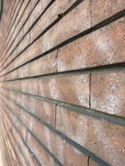 The image shows a close-up, angled view of a weathered brick wall. The bricks are reddish-brown and are laid horizontally with dark gray mortar in between. Some of the bricks have white splotches and a rough texture. The perspective suggests the wall recedes into the distance, creating a sense of depth. There are no people or animals visible in the image. The lighting appears natural, casting subtle shadows that highlight the texture of the bricks and mortar. The scene is devoid of any specific urban or natural landmarks, making it impossible to determine the location within Trieste, Italy, based solely on the provided image.
