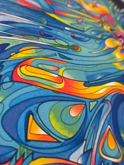 The image shows a close-up view of a colorful, abstract artwork. The dominant colors are shades of blue, yellow, orange, red, green, and white. The composition consists of fluid lines and shapes, creating a sense of movement and depth. Some forms appear to be geometric, like a triangle. The artwork has a textured appearance, possibly indicating it is painted on canvas or another fabric. I cannot find any information related to the location of this work in Novo-Nikol'skoe, Russia.