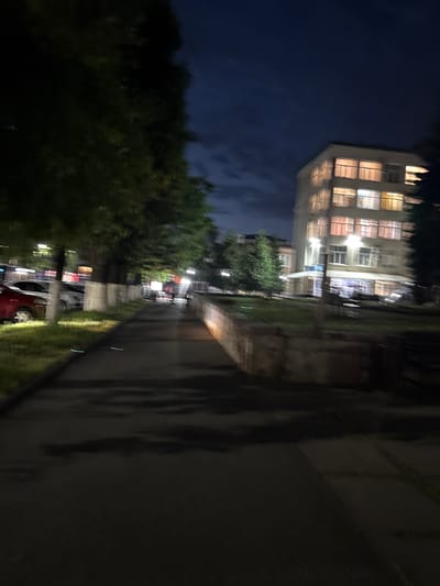 The image depicts a nighttime scene in Vladikavkaz, Russia, likely in a residential or commercial area. On the right side of the frame, a multi-story building with illuminated windows is visible against a dark blue sky. The building appears to be an apartment complex or an office building. In the foreground, a paved walkway runs alongside a grassy area with trees. Several cars are parked along the side of the walkway, with a red car being most prominent. A person is seen walking on the walkway, appearing as a silhouette against the ambient light. The overall impression is one of a quiet evening in the city, with artificial lighting from buildings and streetlights casting shadows.