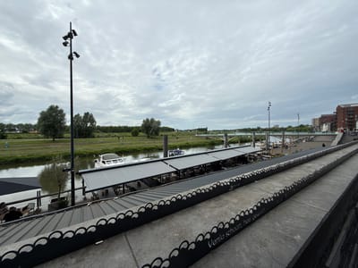 This image presents an elevated, wide-angle view overlooking the Maas river in Venlo, Netherlands, under an overcast sky.
In the immediate foreground, on the right, a multi-tiered concrete or stone promenade features a decorative black metal railing. This railing exhibits a repeating scalloped or wave-like pattern and bears white lettering that reads, from top to bottom, "Original", "Cantus", and "Venlo schoon, heel gewoon".
The mid-ground is dominated by the river, which flows from left to right. Along the river's edge, a series of elongated, covered structures with dark gray awnings or roofs shelter outdoor seating areas, likely for a restaurant or cafe. Tables and chairs are visible beneath these awnings, occupied by several individuals. Moored alongside these structures are multiple boats, including a prominent white cruiser on the left. A small street sign with the word "WERF" is visible near the docks. In the distance, a multi-arched bridge spans the river.
On the far left bank, a verdant, grassy area slopes down to the river, interspersed with several trees. A path runs along the top of this bank, where multiple distant figures can be observed walking or cycling.
Two tall, slender black poles equipped with multiple spotlights punctuate the scene vertically; one is positioned on the far left, and another is closer to the city structures on the mid-right.
The background on the right reveals a segment of the city, characterized by multi-story buildings constructed primarily of brick and concrete.
The sky is uniformly overcast, covered by dense, light gray clouds, indicating diffuse lighting with no distinct shadows. Several people are sparsely distributed across the scene: a few at the riverfront cafe, several on the distant riverbank path, and one individual seated on the steps or promenade in the middle distance towards the bridge.
