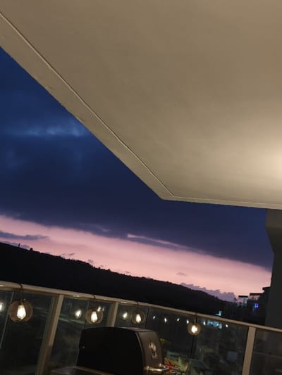 A twilight scene from a balcony captures a pink and purple sunset sky. The horizon reveals a dark silhouette of a hill. Decorative hanging lights illuminate the railing. A black grill occupies the foreground. The upper portion of the image is dominated by a white ceiling structure that blocks a portion of the sky. Tall buildings with lights in the background.