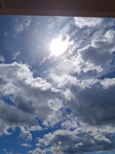 An upward-facing photographic capture taken in Mörkö, Sweden, depicts a bright, partly cloudy sky. The top approximately 10-15% of the frame is occupied by a dark, flat, horizontal architectural element, likely an overhang or roofline, showing minimal texture and a faint reflective highlight on its underside toward the left. Below this structure, the sky presents a gradient of blue hues, from a deeper azure in clearer sections to a brilliant white-blue around the solar disc.

The sun is positioned in the upper right quadrant, appearing as an intensely bright, white light source, partially obscured by clouds. Its presence generates a significant lens flare and radiant light shafts that penetrate the surrounding cloud formations. The sky is populated with a mix of cloud types: large, voluminous cumulus clouds dominate, ranging from brilliant white, sun-illuminated forms to darker, shadow-laden grey masses, indicating depth and varying light absorption. Smaller, wispy cirrus-like clouds are also visible in the clearer blue patches. Extensive areas of clear, vibrant blue sky are discernible between the cloud masses, particularly in the lower left and scattered throughout the mid-frame. No human subjects, animals, or identifiable man-made objects beyond the overhead structure are present in the scene.