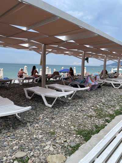 The image depicts a beach scene in Krinitsa, Russia, under a shaded pergola with a light-colored fabric canopy. The foreground is dominated by white plastic beach loungers on a pebble-strewn shore. In the mid-ground, more loungers are occupied by people. Several individuals are seen sunbathing or relaxing. The sea is visible in the background, with a calm surface under a partly cloudy sky. Numerous white beach umbrellas are set up along the shoreline. A few people are in the water, and a small boat is visible further out at sea. The overall atmosphere is one of leisure and recreation.