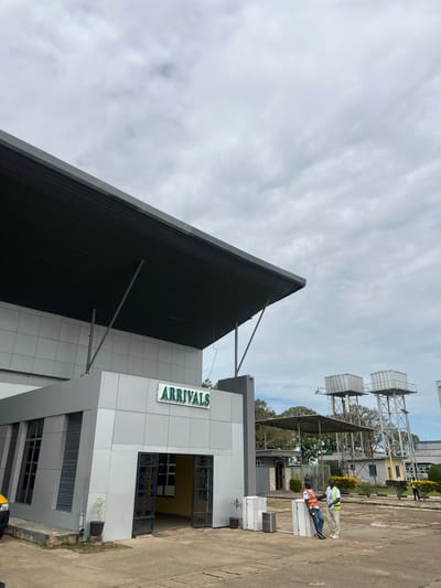 This image captures the "ARRIVALS" section of an airport building in Kwi, Nigeria, under a heavily overcast sky. The primary structure is a modern, grey building with a large, dark grey corrugated roof, supported by grey angular beams. A green sign with white text reading "ARRIVALS" is mounted above the main entrance, which features ornate black metal gates.

In the mid-ground, two men are interacting. One man wears an orange high-visibility vest over a dark shirt, jeans, and sneakers, leaning against a stack of white utility boxes. The second man wears a yellow high-visibility vest over a light shirt, light trousers, and light shoes, standing facing the first man. Further in the background, another person is partially visible near some foliage to the right.

To the right and behind the main building, two tall, grey metal water towers with square tanks dominate the skyline. Several smaller, utilitarian buildings and mature trees are scattered across the background. The foreground consists of a concrete paved area, showing some discoloration and minor vegetation growth. Two small potted plants are visible near the building's entrance and by the utility boxes.